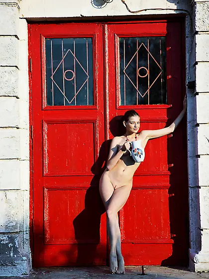 Necklace-wearing naked brunette posing next to a big red door Vi